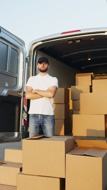 A man wearing a black cap, white t-shirt, and jeans stands with his arms crossed in front of an open van parked outdoors, possibly on a driveway or street, during daylight hours. Inside the van, there are several large cardboard boxes stacked neatly, with some boxes placed on the pavement outside the vehicle. The boxes are taped and appear to be packed for a home relocation, with protective packing materials such as plastic wrap or padding likely used for furniture, though not visible in detail. The scene suggests a furniture transport and packing process related to house removals, coordinated by Man with Van New Barnet, a professional moving company, as part of a comprehensive moving service. The background shows a clear sky and some outdoor environment, emphasizing an active moving operation with items ready for transport to a new property.