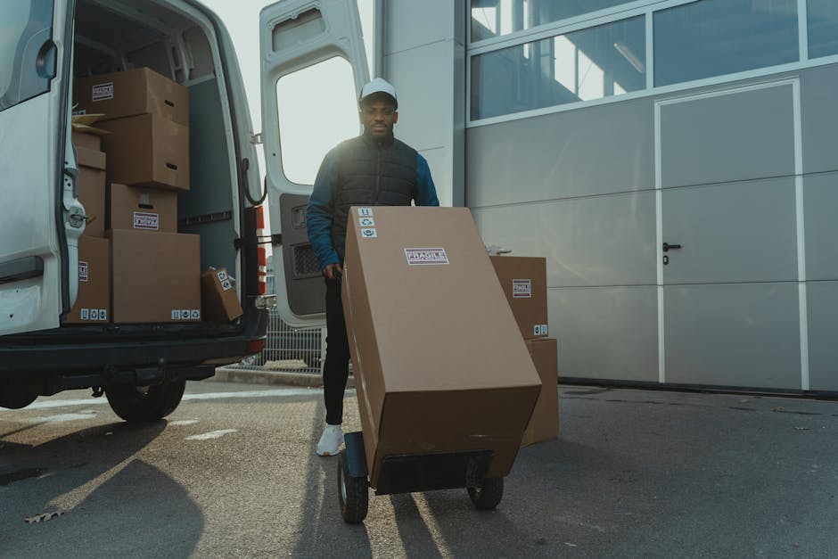 A man with dark skin, wearing a black vest over a blue long-sleeve shirt, black pants, white sneakers, and a white cap, is standing outside a white moving van with open doors at a house relocation site. Inside the van, several brown cardboard boxes of varying sizes are stacked, some with packing labels and tape. The man is holding onto a large, labeled cardboard box positioned on a black dolly with two wheels, which is on the pavement next to the vehicle. The scene is outdoors with natural lighting, near a building with white panel walls and a closed door. The environment reflects a typical furniture transport and packing process involved in home removals, as part of professional relocation services provided by Man with Van New Barnet, supporting customer moves efficiently and securely.