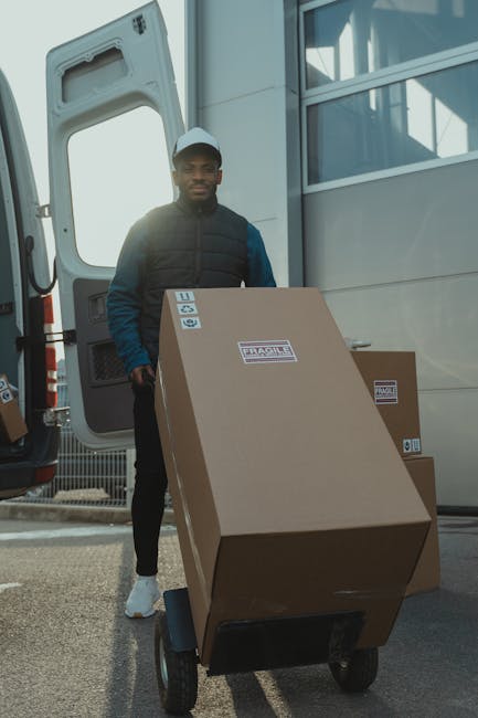 A man wearing a black cap, white t-shirt, and jeans stands with his arms crossed in front of an open van parked outdoors, possibly on a driveway or street, during daylight hours. Inside the van, there are several large cardboard boxes stacked neatly, with some boxes placed on the pavement outside the vehicle. The boxes are taped and appear to be packed for a home relocation, with protective packing materials such as plastic wrap or padding likely used for furniture, though not visible in detail. The scene suggests a furniture transport and packing process related to house removals, coordinated by Man with Van New Barnet, a professional moving company, as part of a comprehensive moving service. The background shows a clear sky and some outdoor environment, emphasizing an active moving operation with items ready for transport to a new property.
