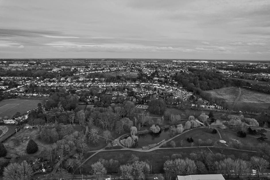 A black and white aerial photograph showing a suburban area with a large park in the foreground, featuring winding pathways, numerous trees with sparse or full foliage, and a small pond. Surrounding the park are residential houses and streets extending into the distance. The background displays a sprawling cityscape with densely packed buildings and open fields under a cloudy sky. This image captures the environment where home relocation and furniture transport services by Man with Van New Barnet may be carried out, illustrating the typical urban and suburban landscape involved in moving logistics between Oakleigh Park and High Barnet.